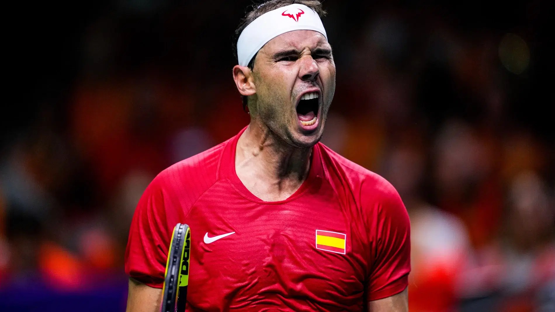 Tennis player in a red shirt with a Spain flag patch shouting on the court, white headband visible.